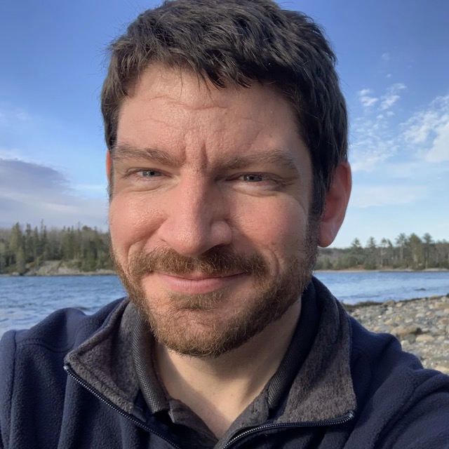 Justin Russell smiling while siting on a pebble beach in front of a body of water